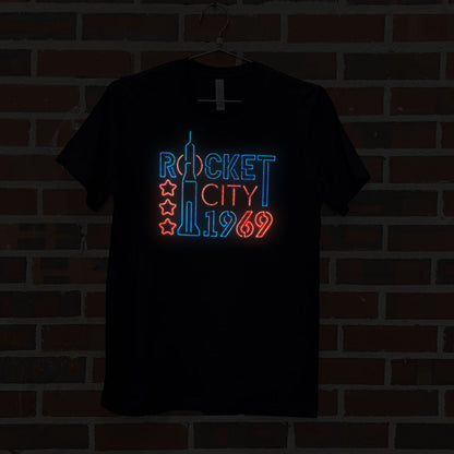 Image of neon lights design with text "Rocket City 1969" on black t-shirt hanging against brick wall at Lowe Mill