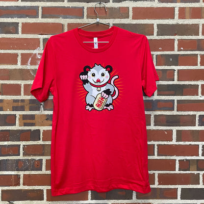 Thumbnail of a possum in the "lucky cat" pose on a red shirt against a red brick wall
