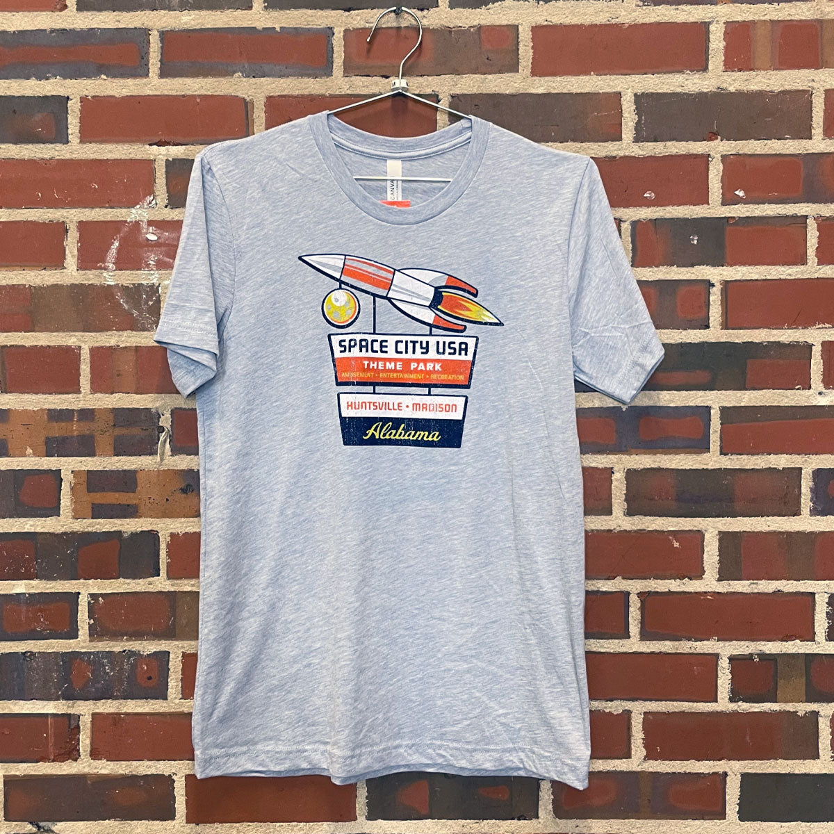 Image of fun retro Space City USA theme park design on light heather blue t-shirt hanging against brick wall at Lowe Mill