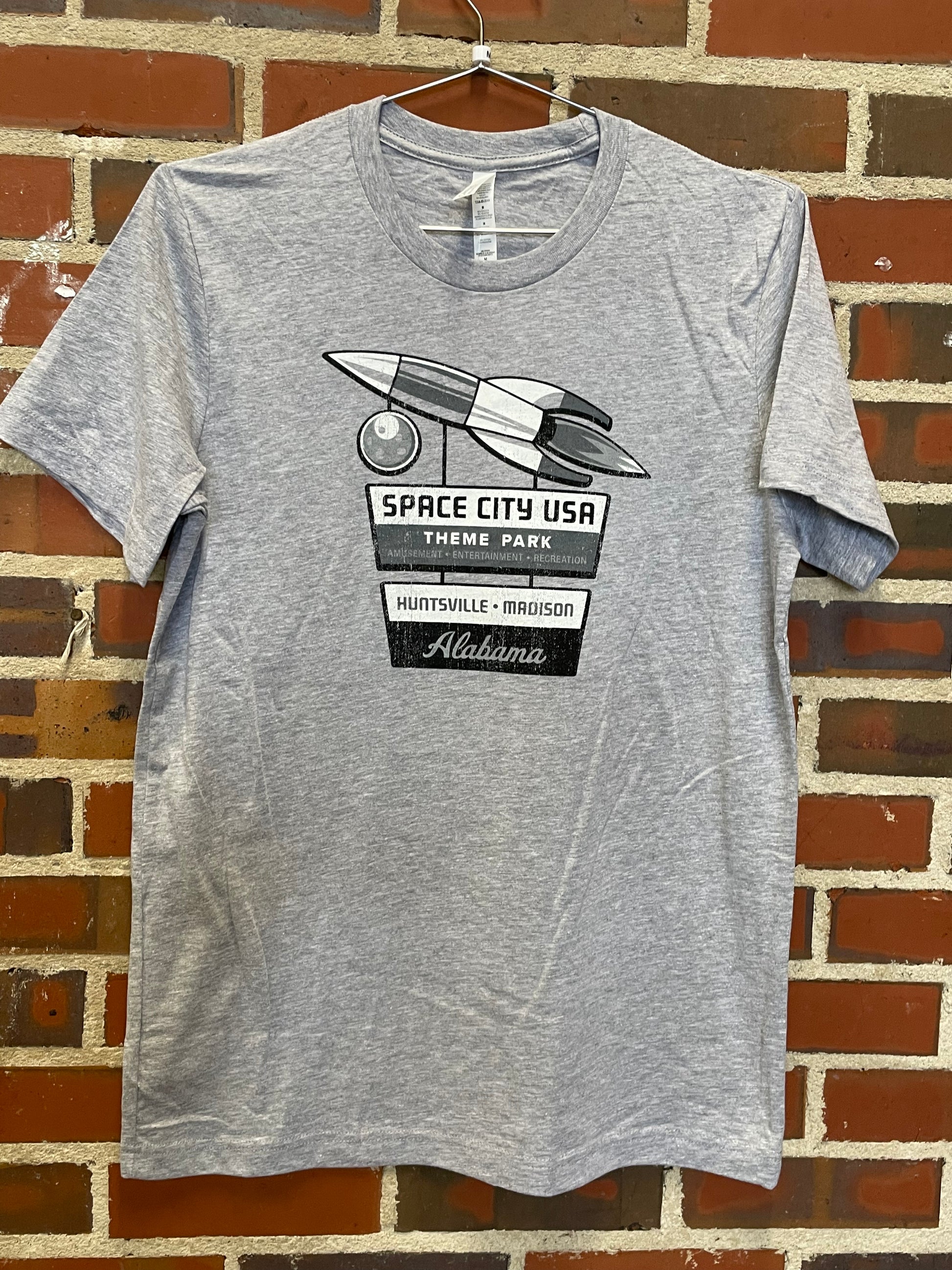 Image of fun retro Space City USA theme park design on light heather grey t-shirt hanging against brick wall at Lowe Mill