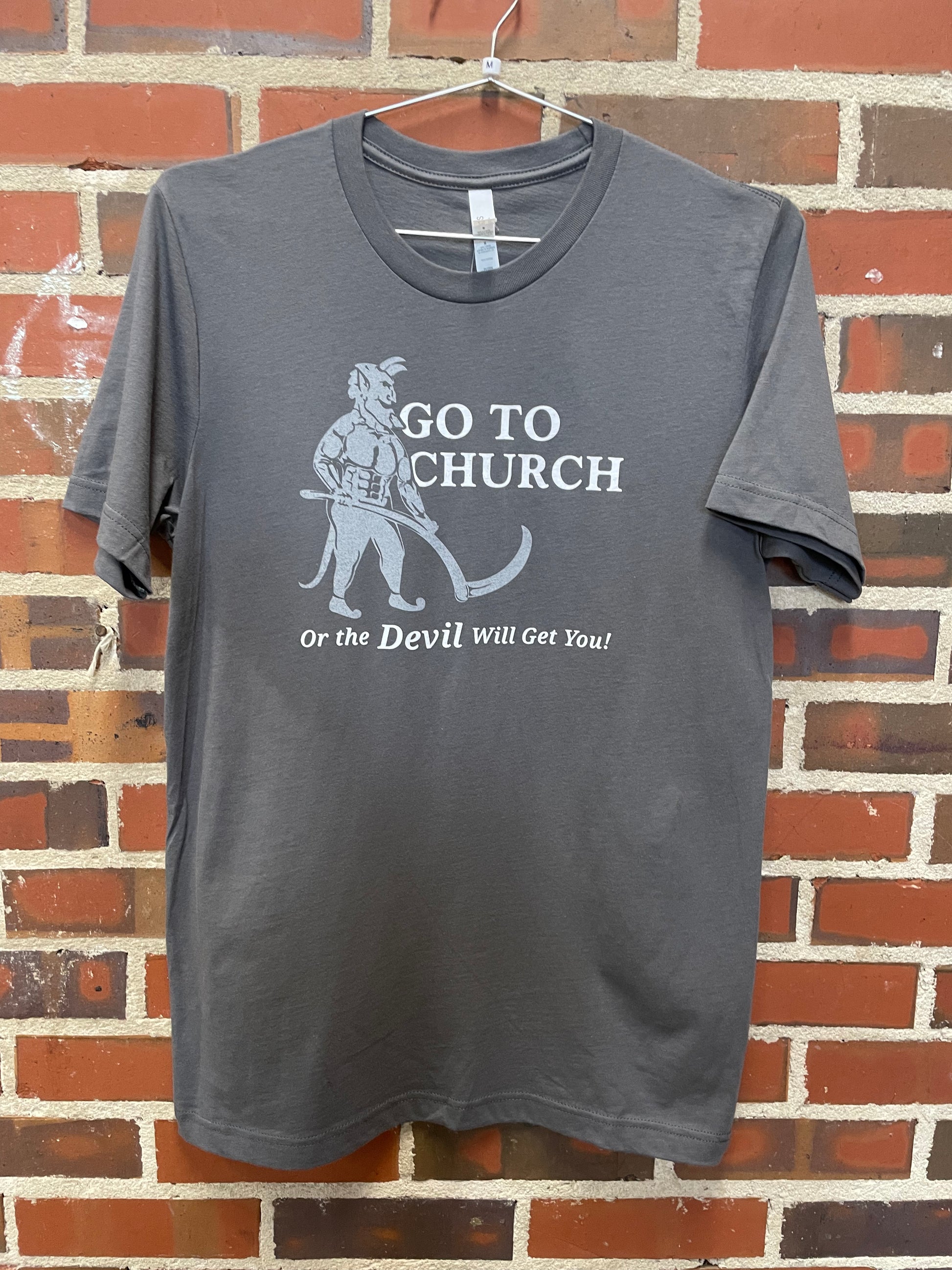 Image of a devil with text " Go to Church or the Devil Will Get You!" on grey t-shirt hanging against brick wall in Lowe Mill