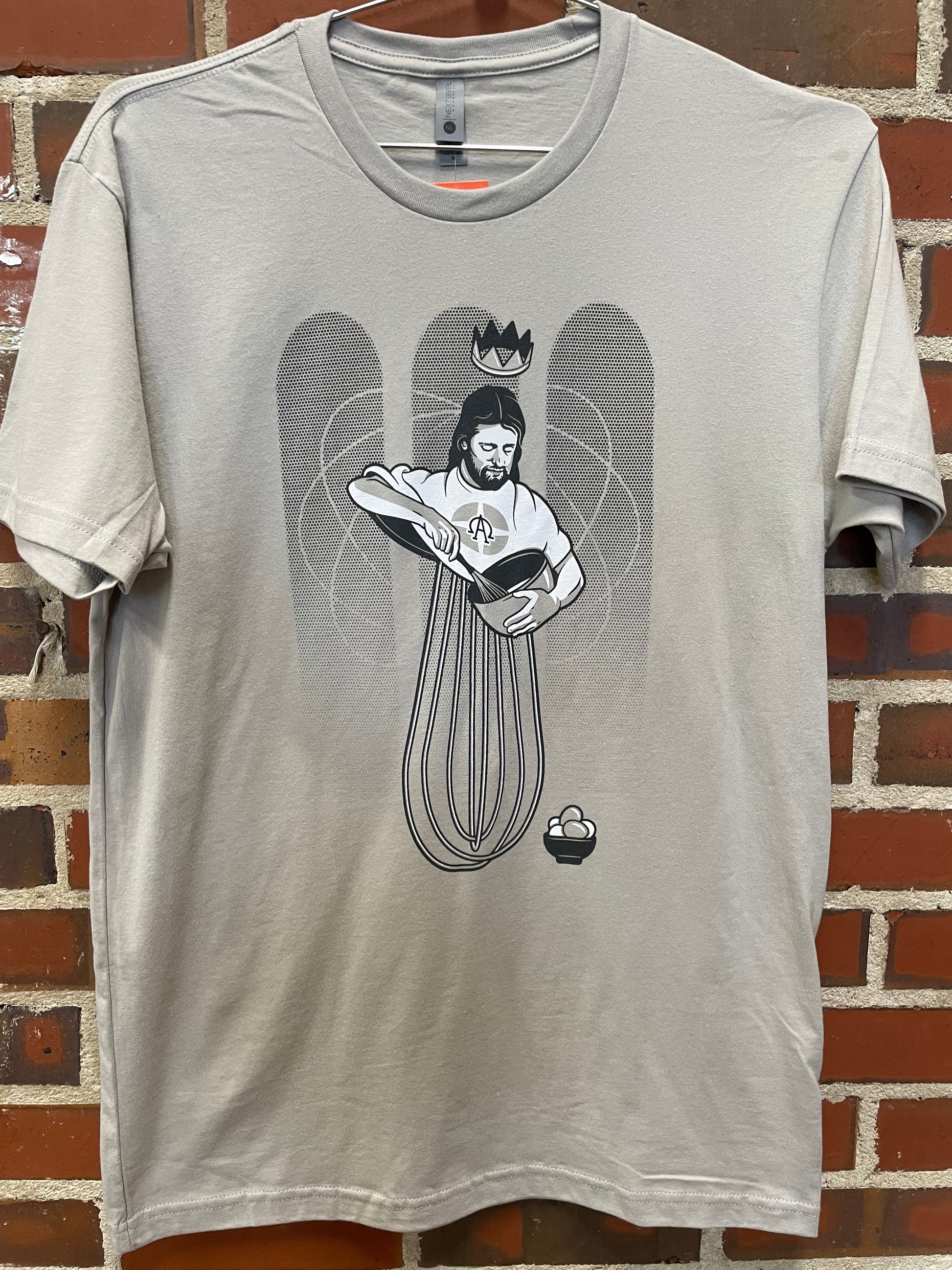 Image of Eggbeater Jesus with mixing bowl on grey t-shirt hanging against brick wall in Lowe Mill