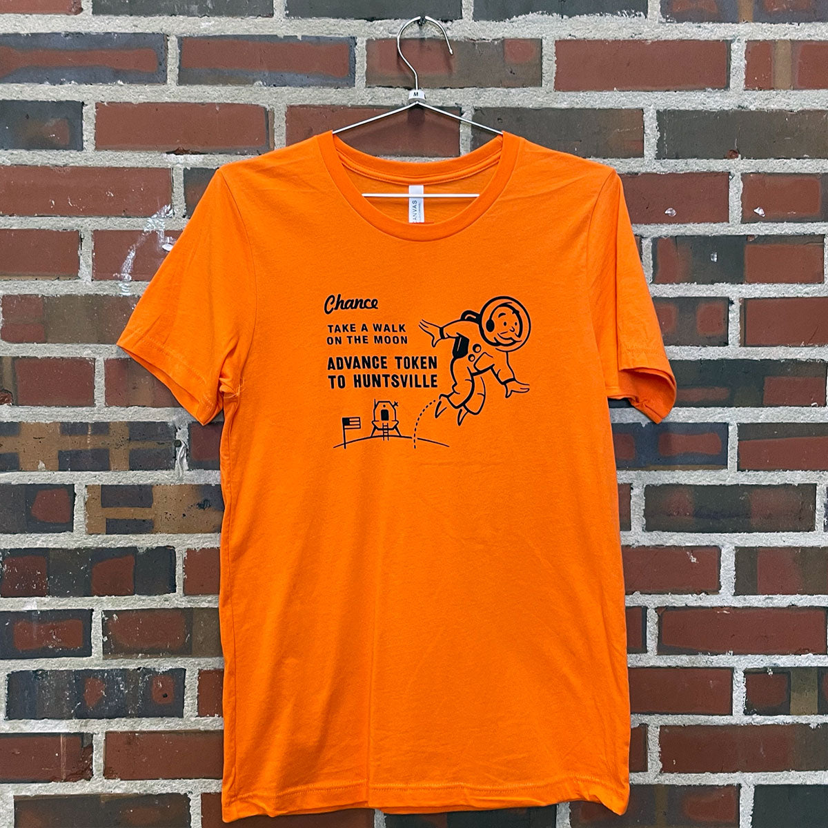 Image of Monopoly man on the moon with text " Chance; Take a walk on the moon; Advance token to Huntsville" on orange t-shirt hanging against brick wall at Lowe Mill