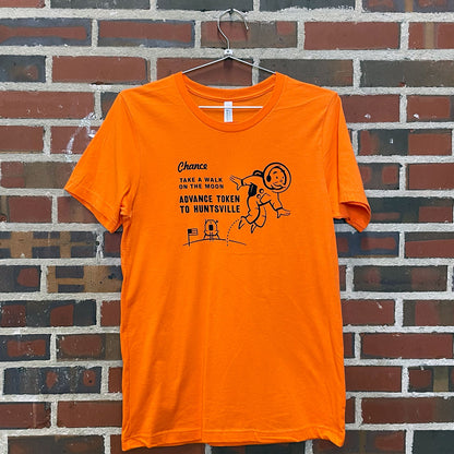 Image of Monopoly man on the moon with text " Chance; Take a walk on the moon; Advance token to Huntsville" on orange t-shirt hanging against brick wall at Lowe Mill