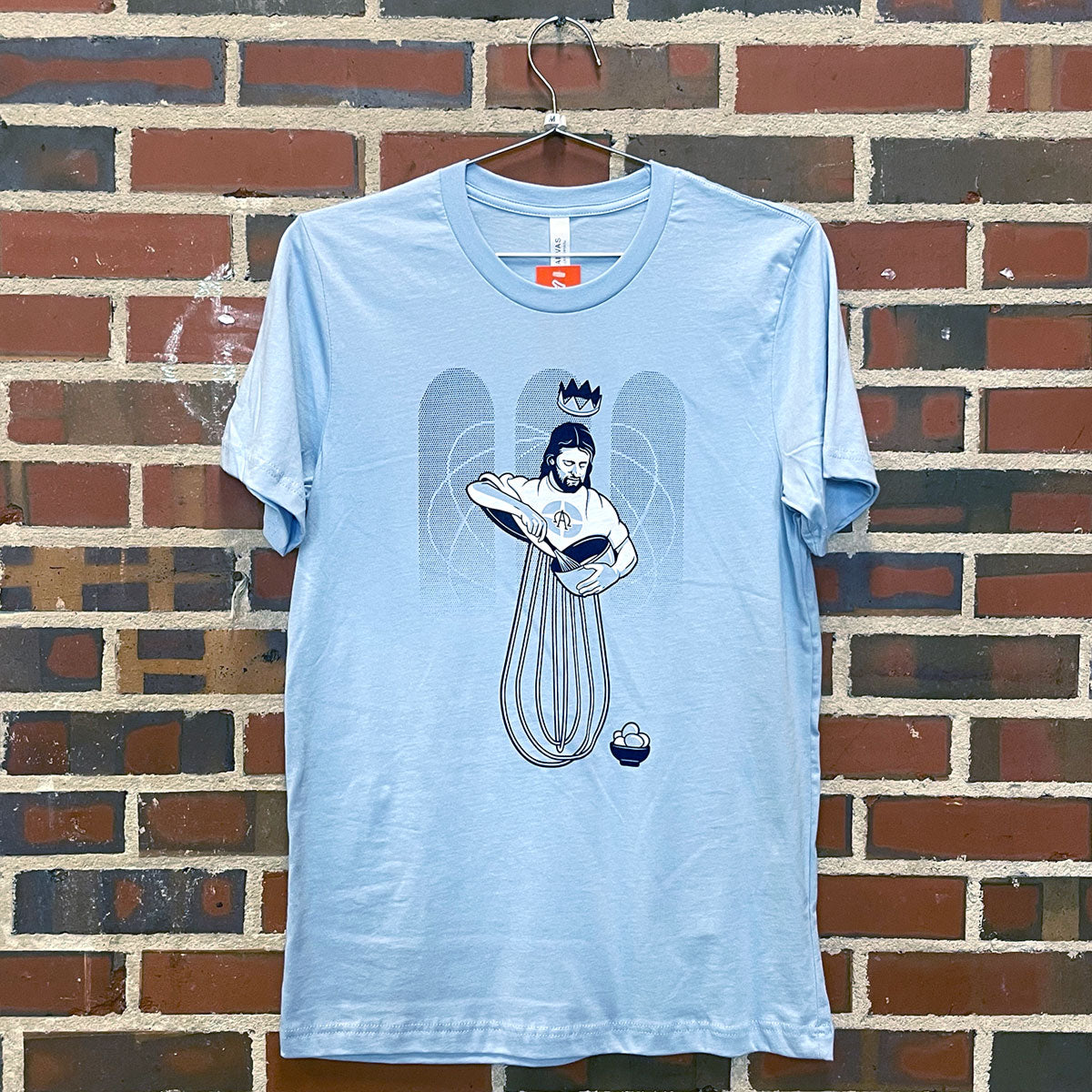 Image of Eggbeater Jesus with mixing bowl on blue t-shirt hanging against brick wall at Lowe Mill