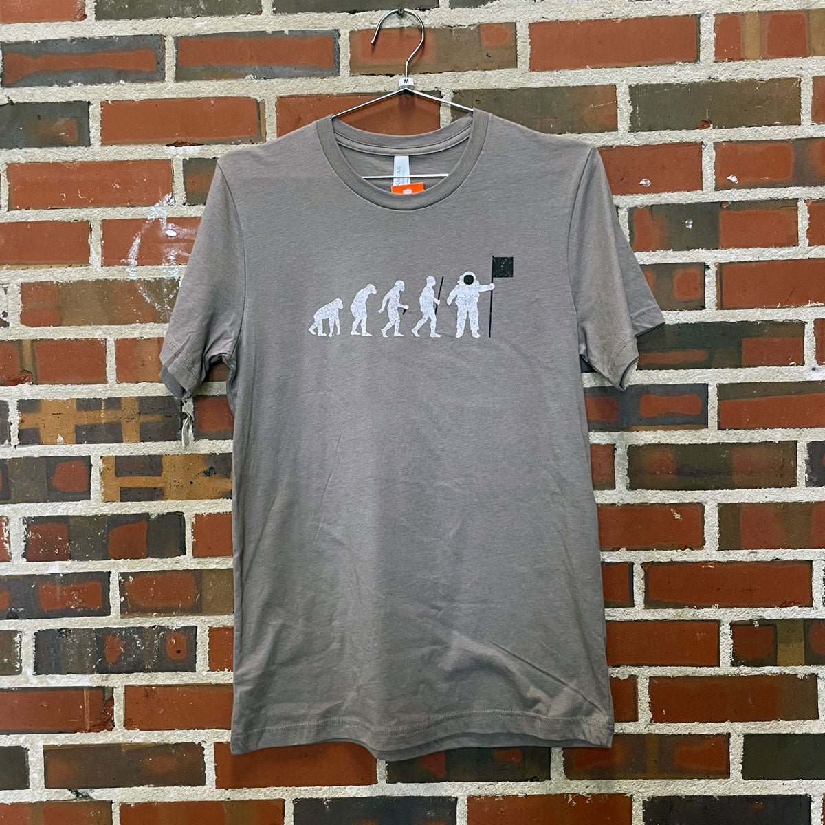 Image of evolution progression from ape to spaceman on grey t-shirt hanging against brick wall at Lowe Mill