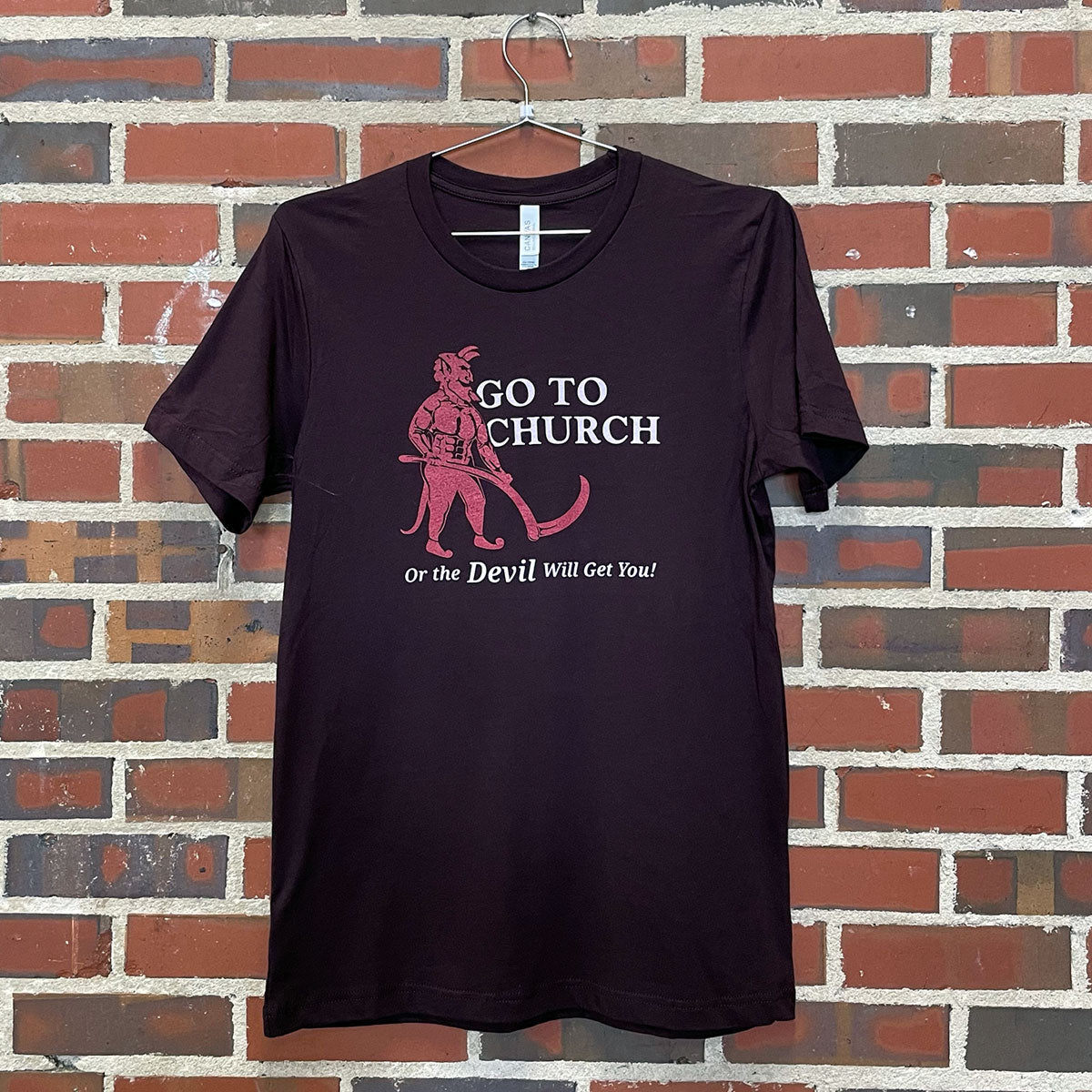 Image of a red devil with text " Go to Church or the Devil Will Get You!" on dark t-shirt hanging against brick wall at Lowe Mill