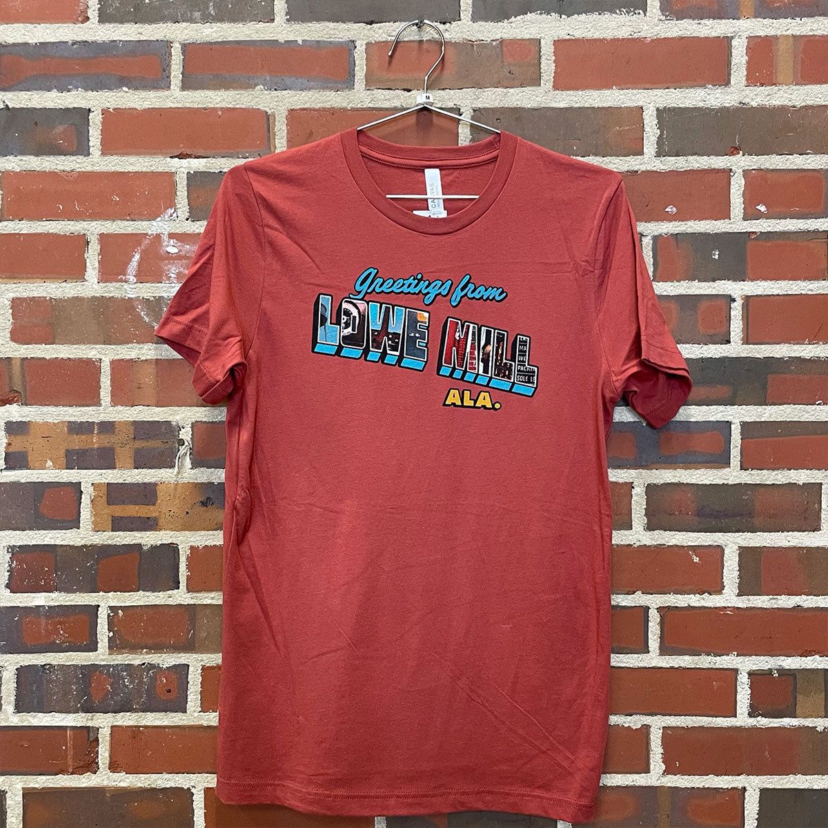 Image of Greetings from Lowe Mill design on red t-shirt hanging against brick wall at Lowe Mill