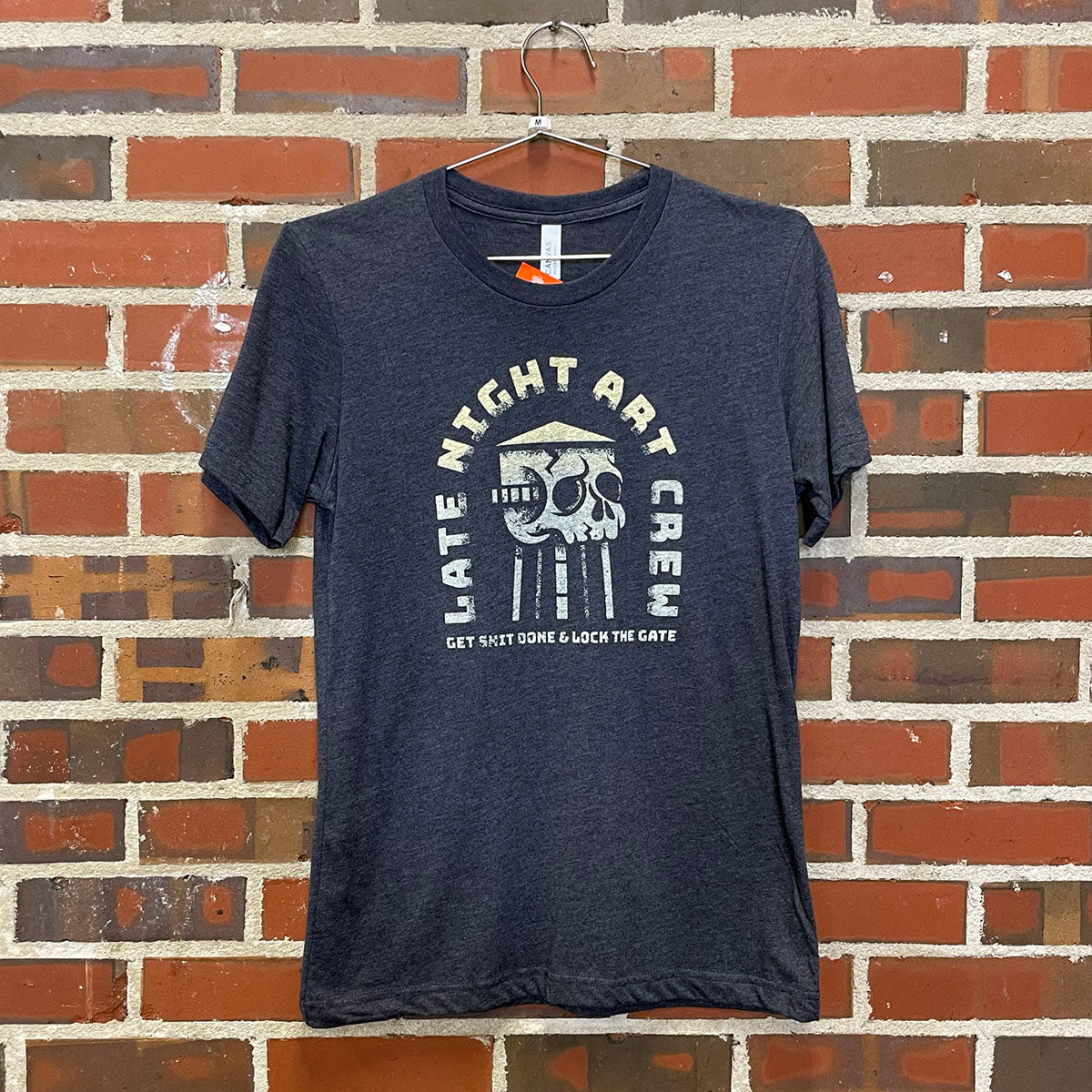 Thumbnail of text "late night art crew" with a skull shaped watertower on a dark blue shirt against a red brick wall