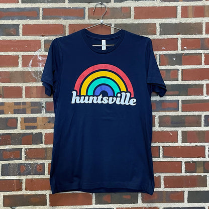 Blue T-Shirt of the text "huntsville" with a rainbow over it hanging against a red brick wall